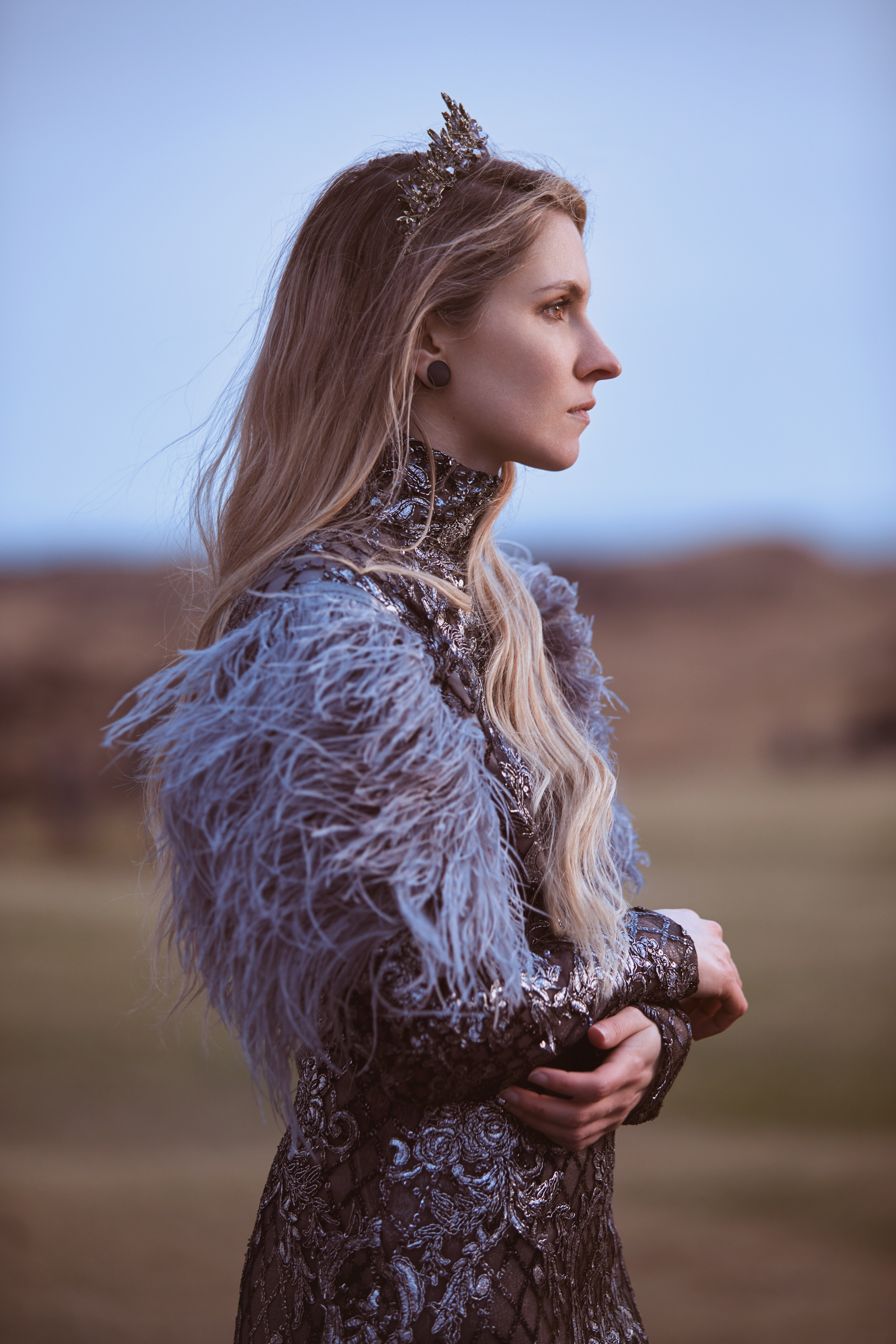 Side profile of an elegant woman with a crystal crown, shimmering feathered dress, against a soft blue sky and blurred landscape.