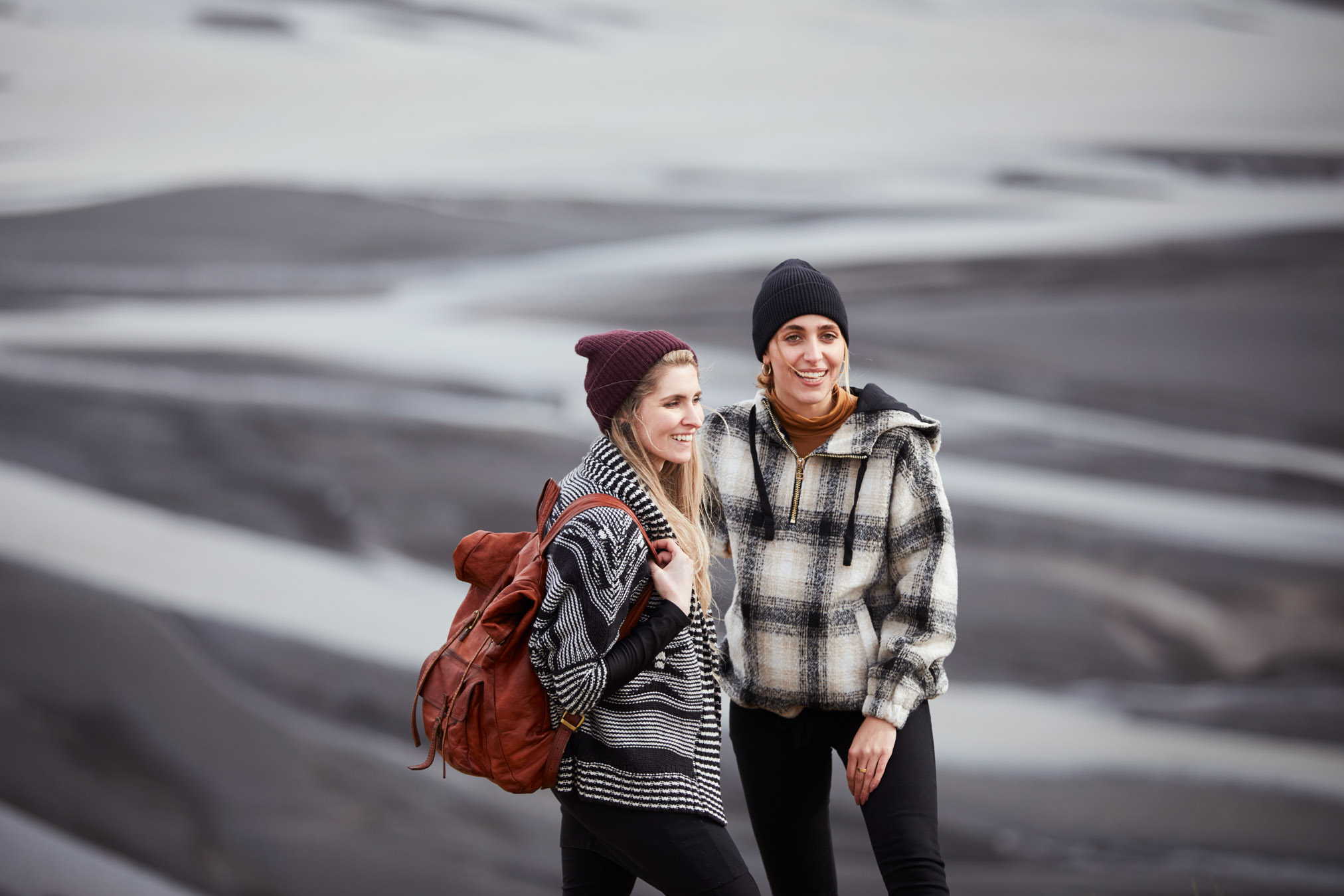 Outdoor portrait of two smiling women in beanies and warm jackets. Blurred background reveals striking light and dark patterns.