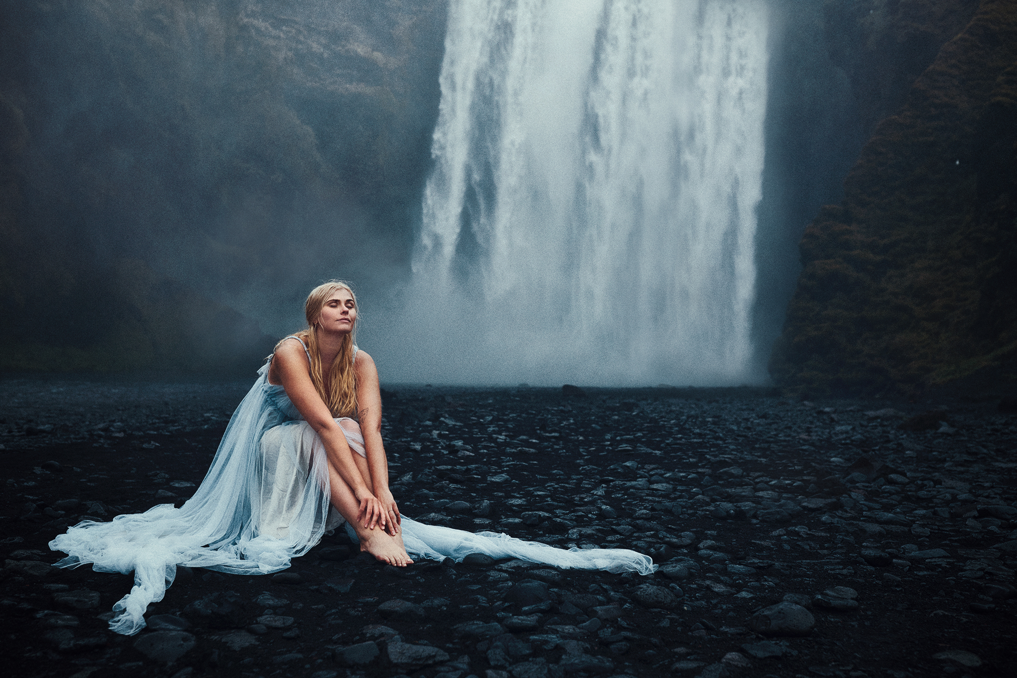 Ethereal, moody portrait: Blonde woman in flowing dress sits peacefully on dark rocks, eyes closed, before a powerful, misty waterfall.
