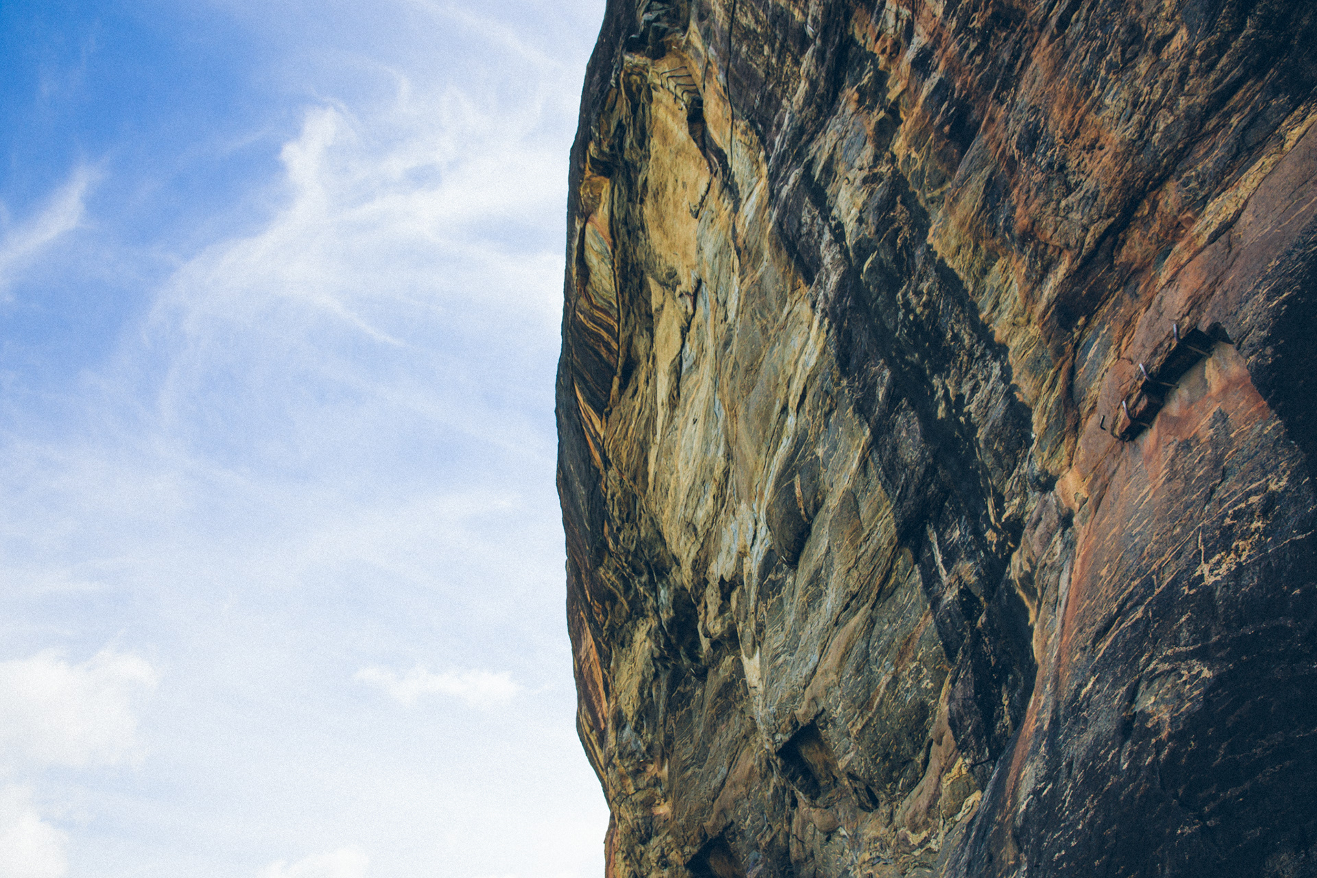 Textured rock face, rich with natural patterns and warm hues, looms against a vibrant blue sky with soft, scattered clouds. A hint of human ascent.